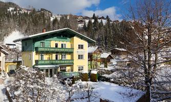 A beautiful yellow house with a green balcony in a snowy landscape. Surrounded by trees and mountains under a clear blue sky.