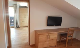 A cozy room with wooden flooring and a desk. In the background, there is a television on the wall and access to another room.