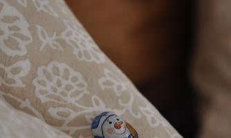 A small snowman figure with a blue scarf lies on a patterned pillow. The background is blurry and shows wooden surfaces.