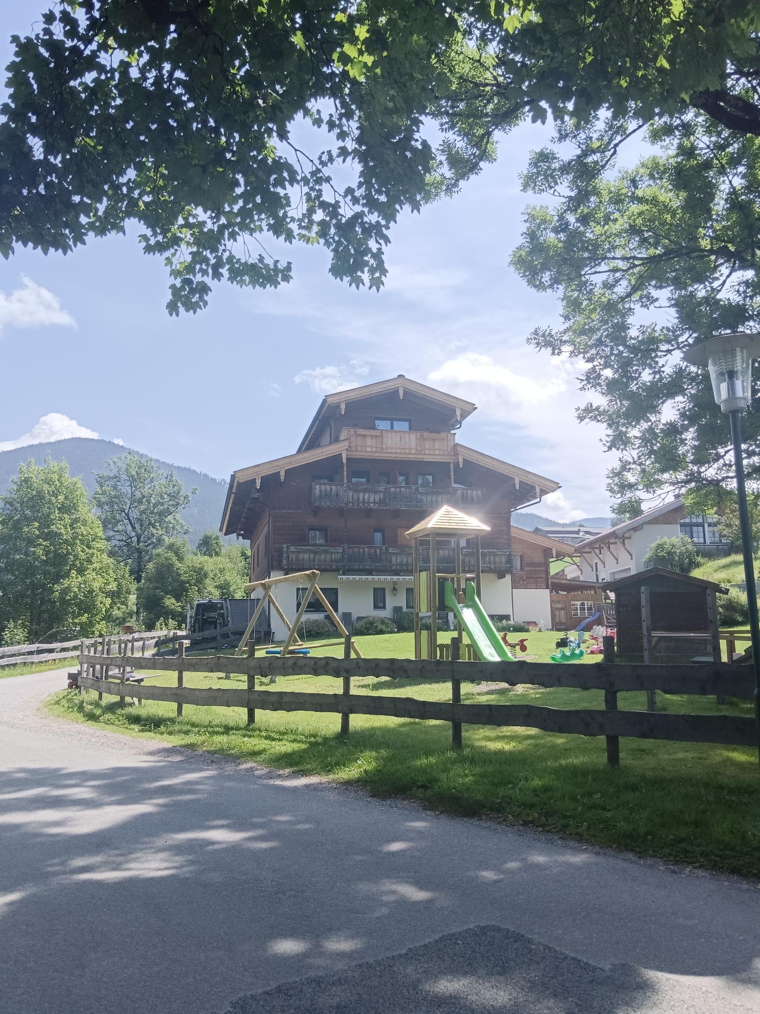 A large wooden house surrounded by trees and meadows. In the foreground, there is a playground with a slide and swings.