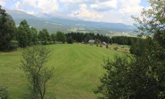 A green meadow with trees and hills. In the background, mountains and a blue sky can be seen.