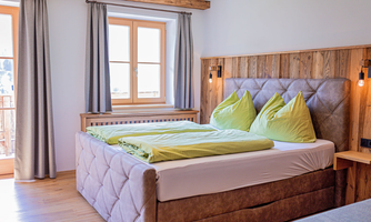 A modern bedroom with a large bed and cozy, green cushions. Windows with curtains provide daylight and a welcoming atmosphere.