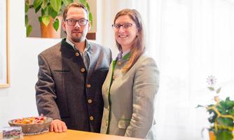 A couple stands at the reception, both wearing traditional Austrian attire. The environment is bright and friendly with green plants in the background.