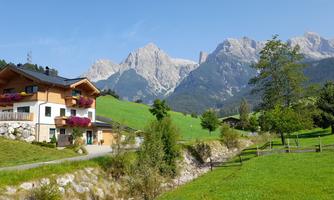 A picturesque house in a green landscape with mountains in the background. The sky is clear and sunny.