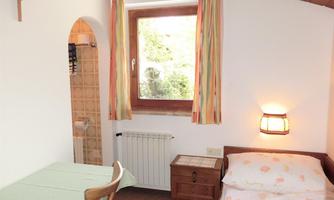 A simple room with a single bed, a table, and a window with colorful curtains. In the background, there is a shower and a radiator.
