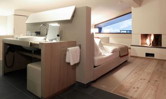 A modern room with a cozy bed and a fireplace. There is a stylish sink and large windows that let in plenty of light.