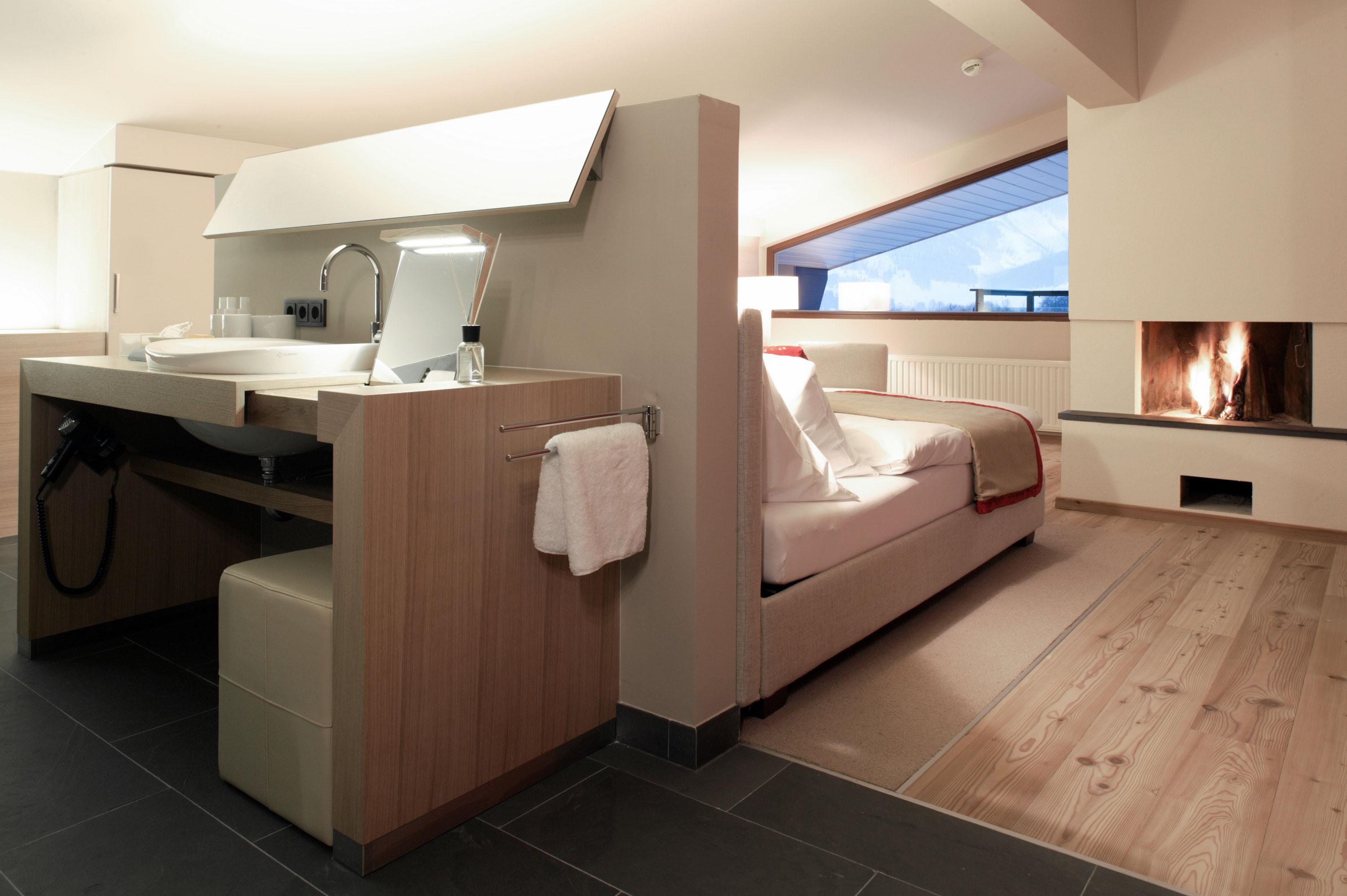 A modern room with a cozy bed and a fireplace. There is a stylish sink and large windows that let in plenty of light.