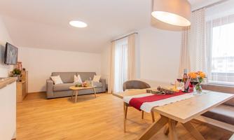 A bright, modern living space with a grey sofa and a dining table. Large windows provide plenty of natural light.