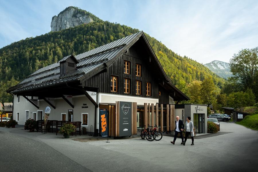 A traditional wooden house in a picturesque setting. In the foreground, two people are passing by and there are bicycles parked.
