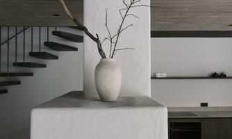 A modern room with a white column pillar and a vase containing a dry branch. In the background, a staircase and a minimalist kitchenette are visible.
