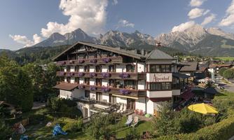A beautiful hotel with flower boxes and balconies amidst a picturesque mountain landscape. In the foreground, there is a well-maintained garden with play opportunities.