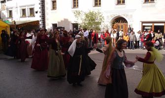 A group of people in traditional clothing is dancing on a festive street. In the background, there are spectators and historical buildings visible.