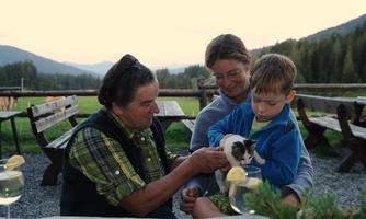 A woman is sitting outside with a child and another woman. They are holding a cat in their hands, while nature is visible in the background.