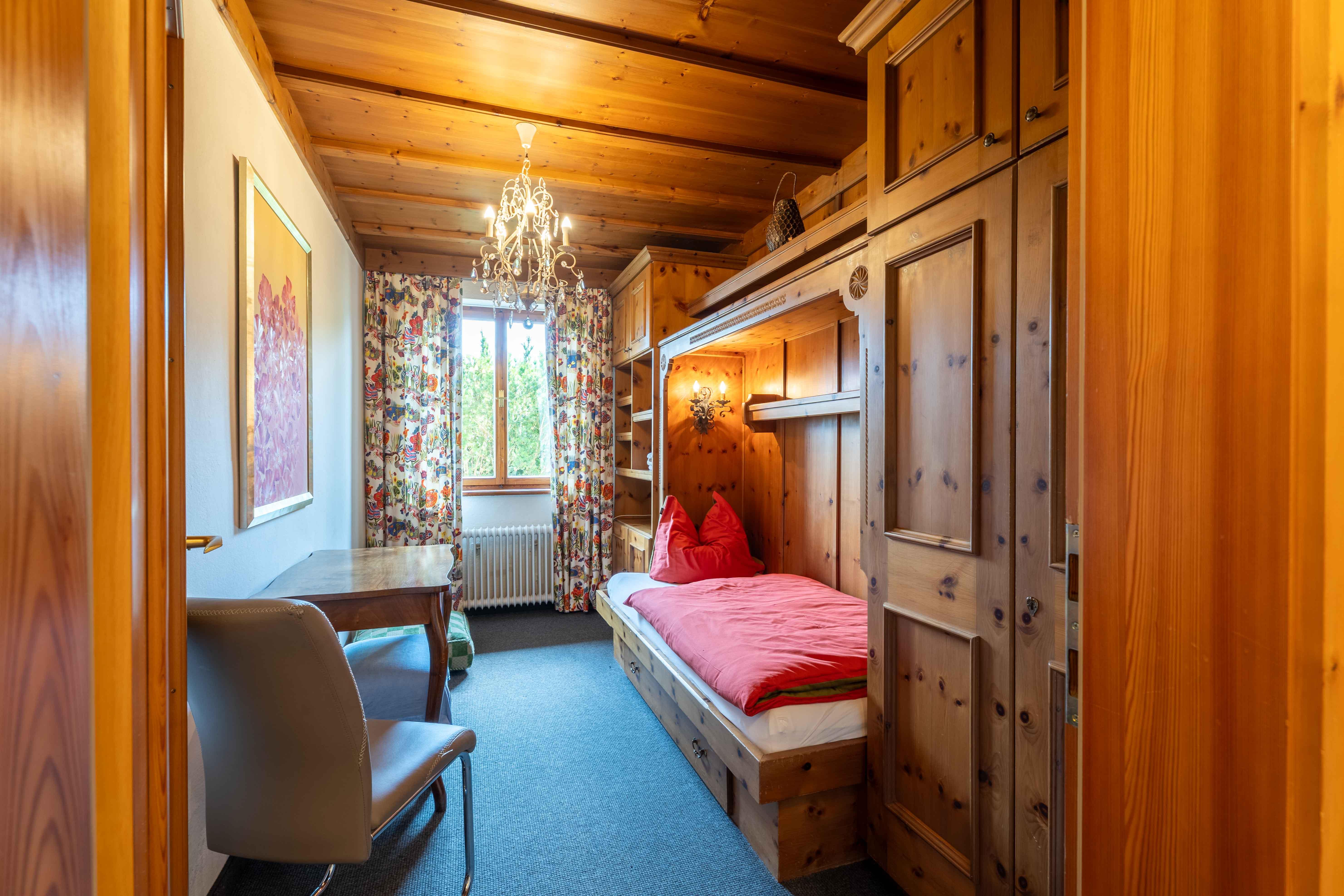 A cozy room with wooden paneling and a single bed. The room is bright and has a window with colorful curtains.