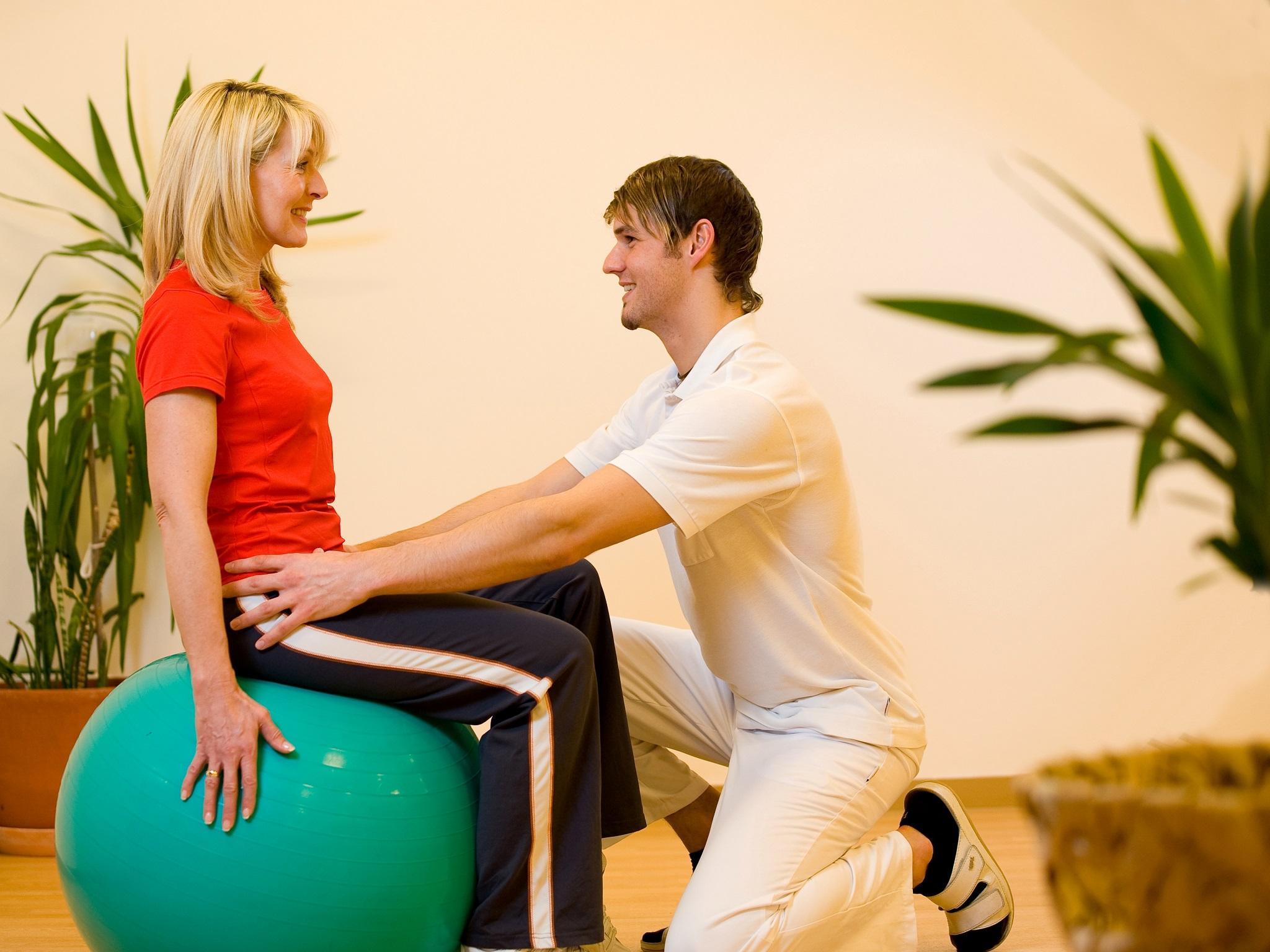 A woman is sitting on a gym ball while a man is helping her. The scene takes place in a bright room with plants.