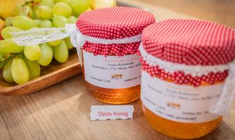 Two jars of honey with red caps and labels are sitting on a wooden table. In the background, green grapes are visible.