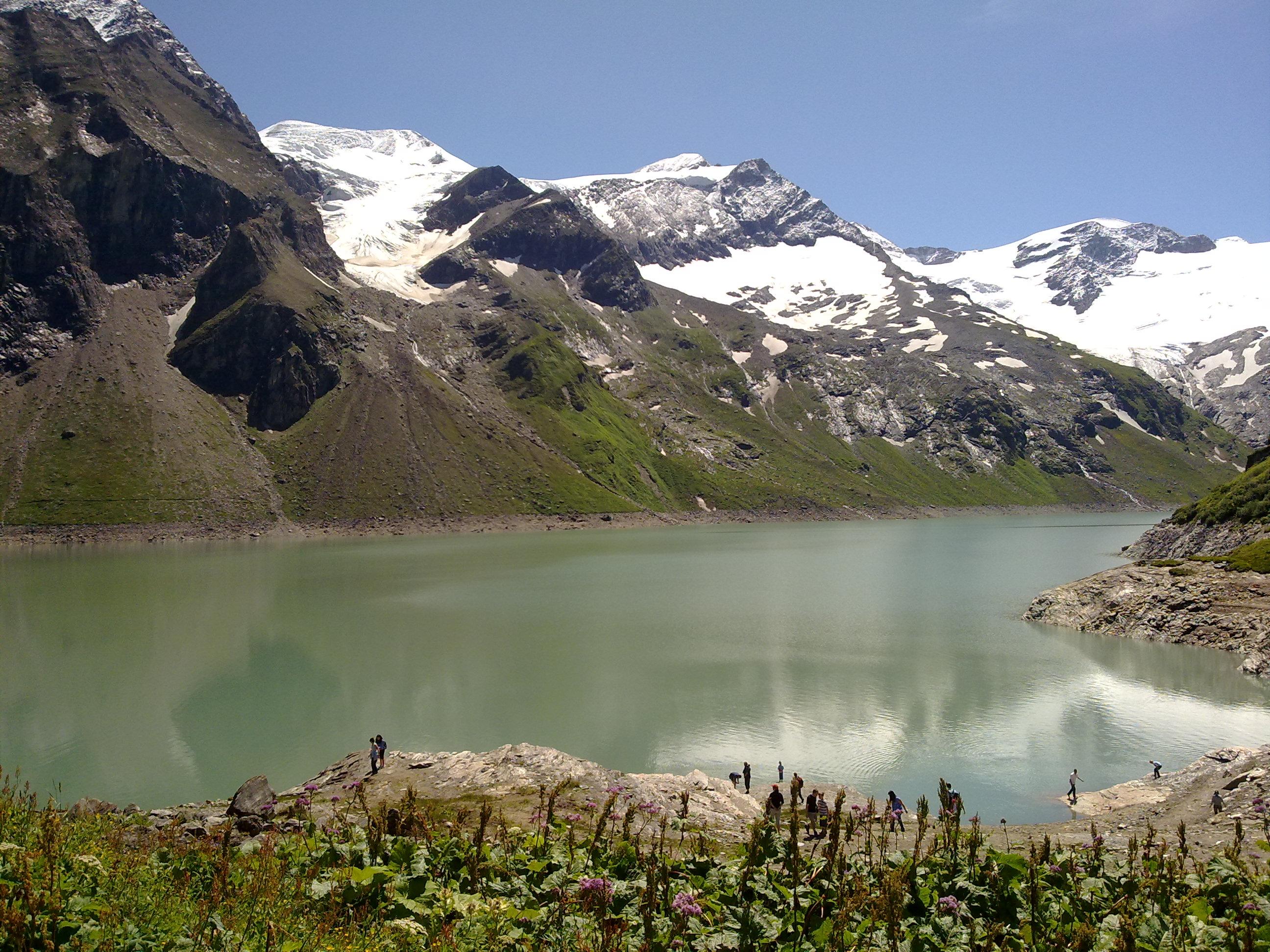 A tranquil lake surrounded by majestic mountains with snow-capped peaks. The sky is clear and the landscape radiates natural beauty.