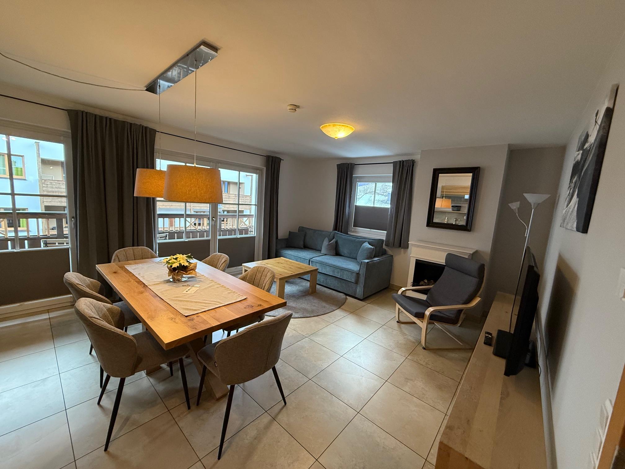 A modern living room with a large dining table and a cozy seating area. The room has large windows and is bright and inviting.