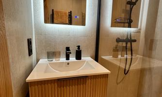 A modern bathroom with a wooden vanity and two illuminated mirrors. The shower is elegantly designed, and the walls are bright and simple.