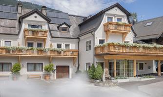A modern building with wooden cladding and balconies. The entrance area features a beautiful garden with green plants.