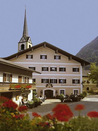 A charming building in alpine style with a tall church tower. Blooming flowers adorn the courtyard and the surroundings are surrounded by mountains.