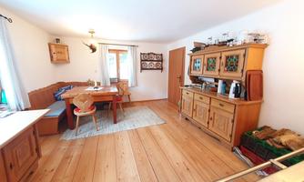 A cozy kitchen with a wooden floor and a dining table. The walls are light and there are traditional furniture pieces as well as windows looking outside.