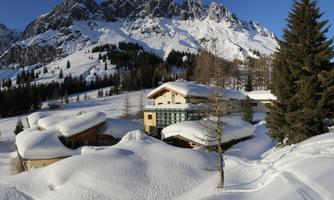 A picturesque winter landscape with snow-covered hills and mountains in the background. In the center, several buildings stand gently enveloped by snow.