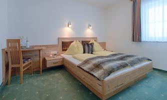 A cozy bedroom with a double bed and light bedding. There is a small table and chairs as well as a window with curtains.