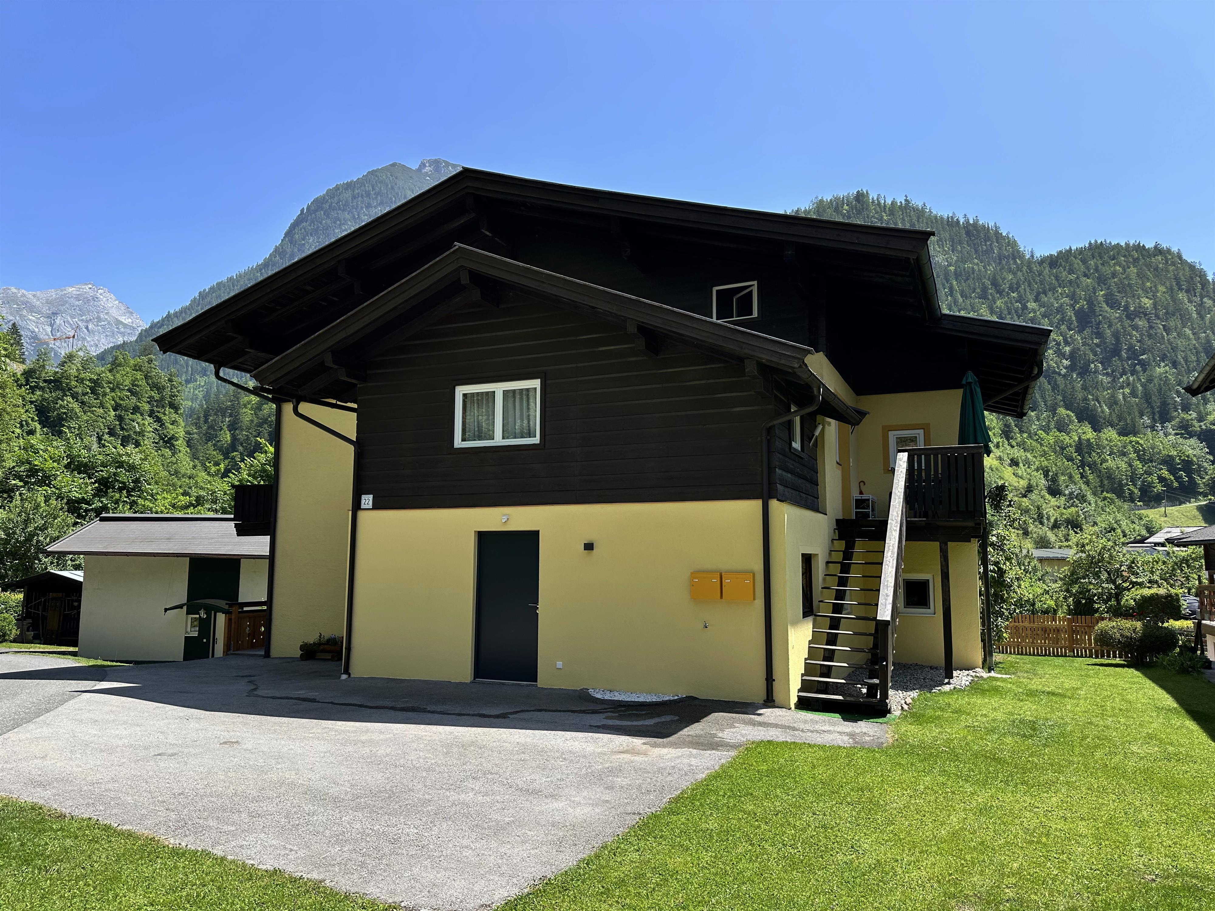 A modern, two-story house with a yellow and dark wood facade. In the background, green mountains and a blue sky are visible.