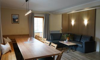 A cozy living space with a large wooden table and a comfortable couch. Soft curtains and warm light sources create an inviting atmosphere.