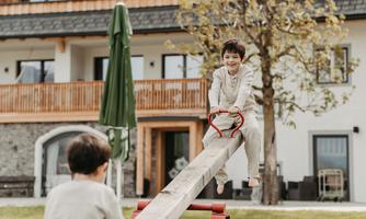 Two children are playing on a seesaw in a garden. In the background, a building with a balcony and trees can be seen.