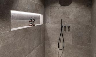 A modern shower with gray tiles and minimalist furnishings. In the background, two bottles are placed on a illuminated shelf.