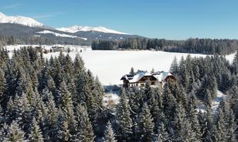 A picturesque winter landscape with snow-covered trees and a solitary house. In the background, snow-covered mountains can be seen under a clear blue sky.