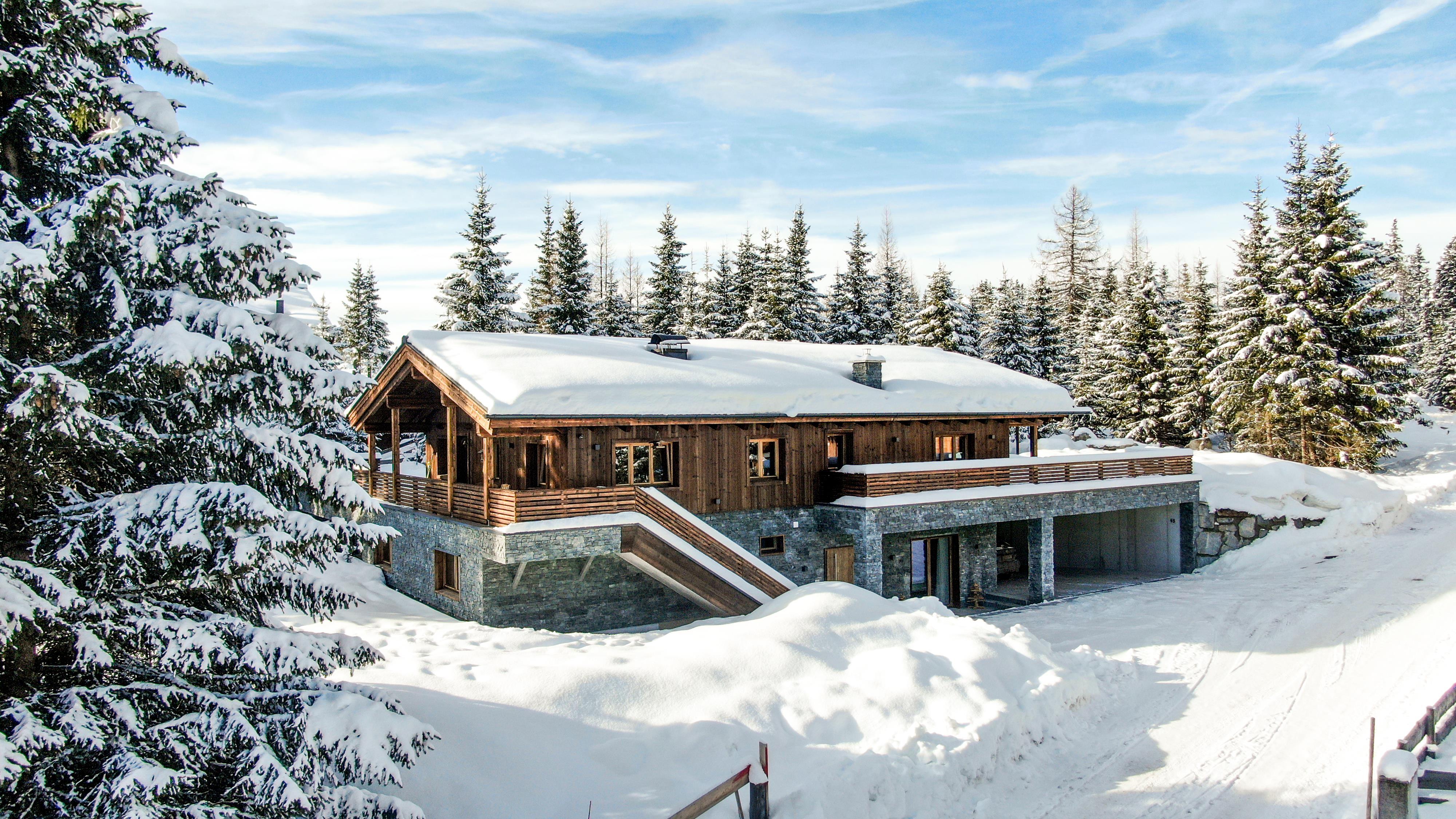 A cozy wooden house in the snow, surrounded by fir trees. The sky is clear and blue, creating a peaceful winter landscape.