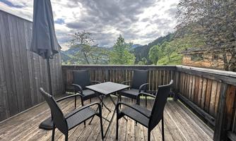 A cozy balcony with a square table and four chairs. In the background, trees and mountains can be seen under a cloudy sky.