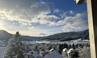 A winter landscape with snow-covered hills and houses. The sky is clear with some clouds, and the sun shines warmly down.