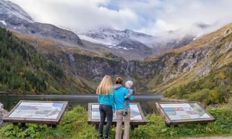 A mother with her child stands by the shore of a clear lake, looking at majestic mountains. Information boards are visible in the foreground.