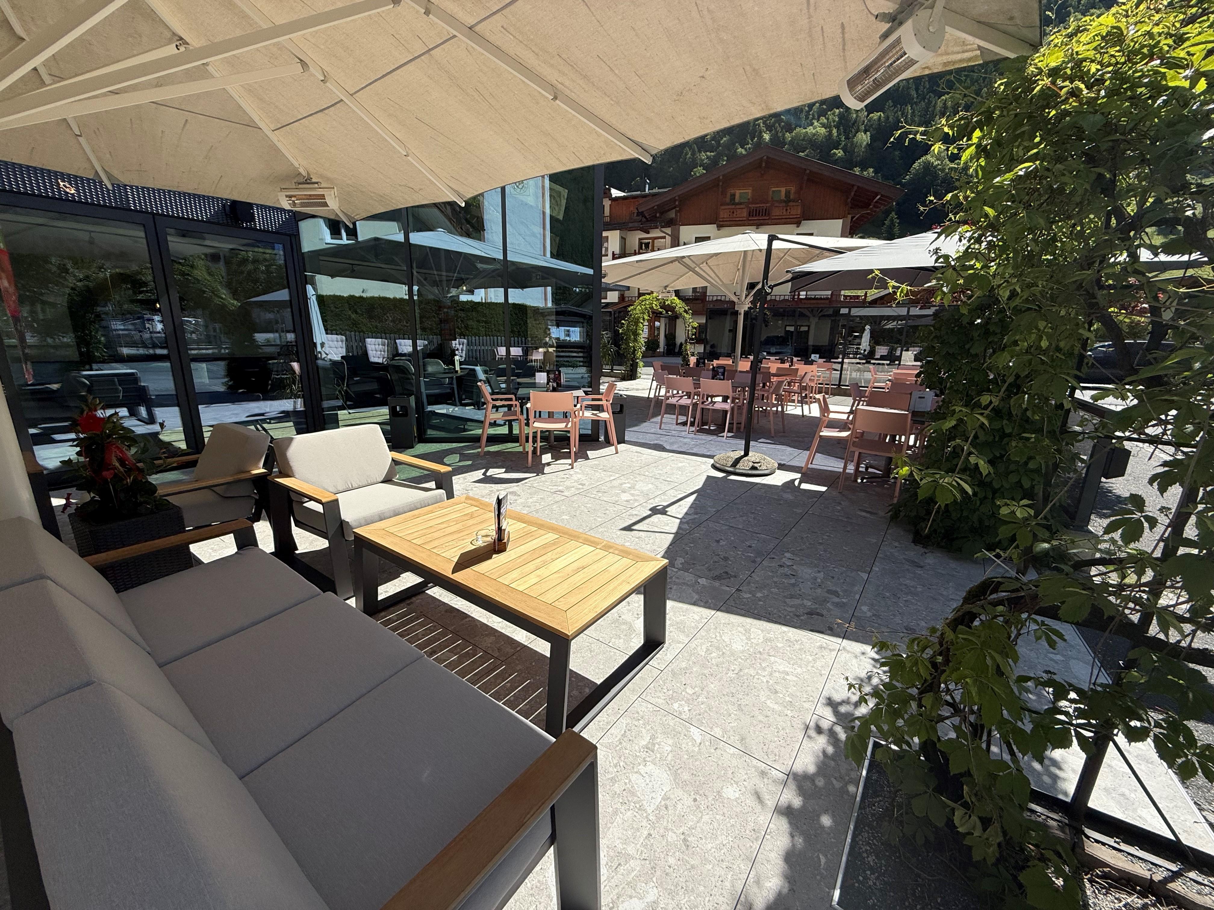 A cozy outdoor terrace with wooden chairs and tables. Umbrellas provide shade, surrounded by plants and a modern atmosphere.