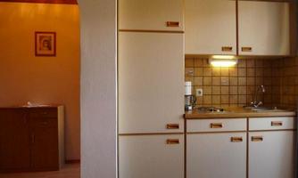 A small, bright kitchen with light cabinets and a small cooking area. In the background, a dining area is visible.