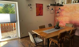 A bright dining room with a large table and wooden chairs. Artworks hang on the wall and there are windows that let in plenty of light.