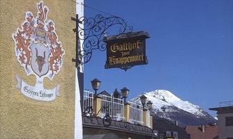 A traditional inn with a decorative sign and a coat of arms on the wall. Snow-capped mountains can be seen in the background.