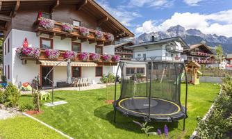 A cozy house with flower boxes and a large garden. In the garden, there is a trampoline, and in the background, mountains can be seen.