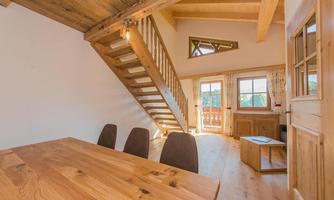 An inviting living and dining room with warm wooden elements. A staircase leads to the upper floor, while large windows provide plenty of natural light.