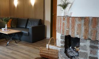 A cozy living area with a black sofa and a wooden table. Next to the sofa stands a traditional fireplace with stones and ash.