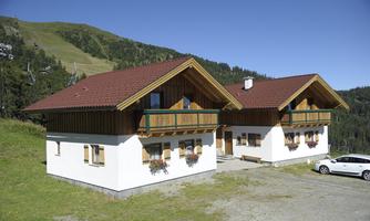 Two charming wooden Alpine chalets with red roofs in a green mountain landscape. The mountains are visible in the background and the weather is sunny.