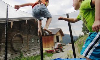 Two children are happily jumping on a trampoline. In the background, a wooden house and nature can be seen.