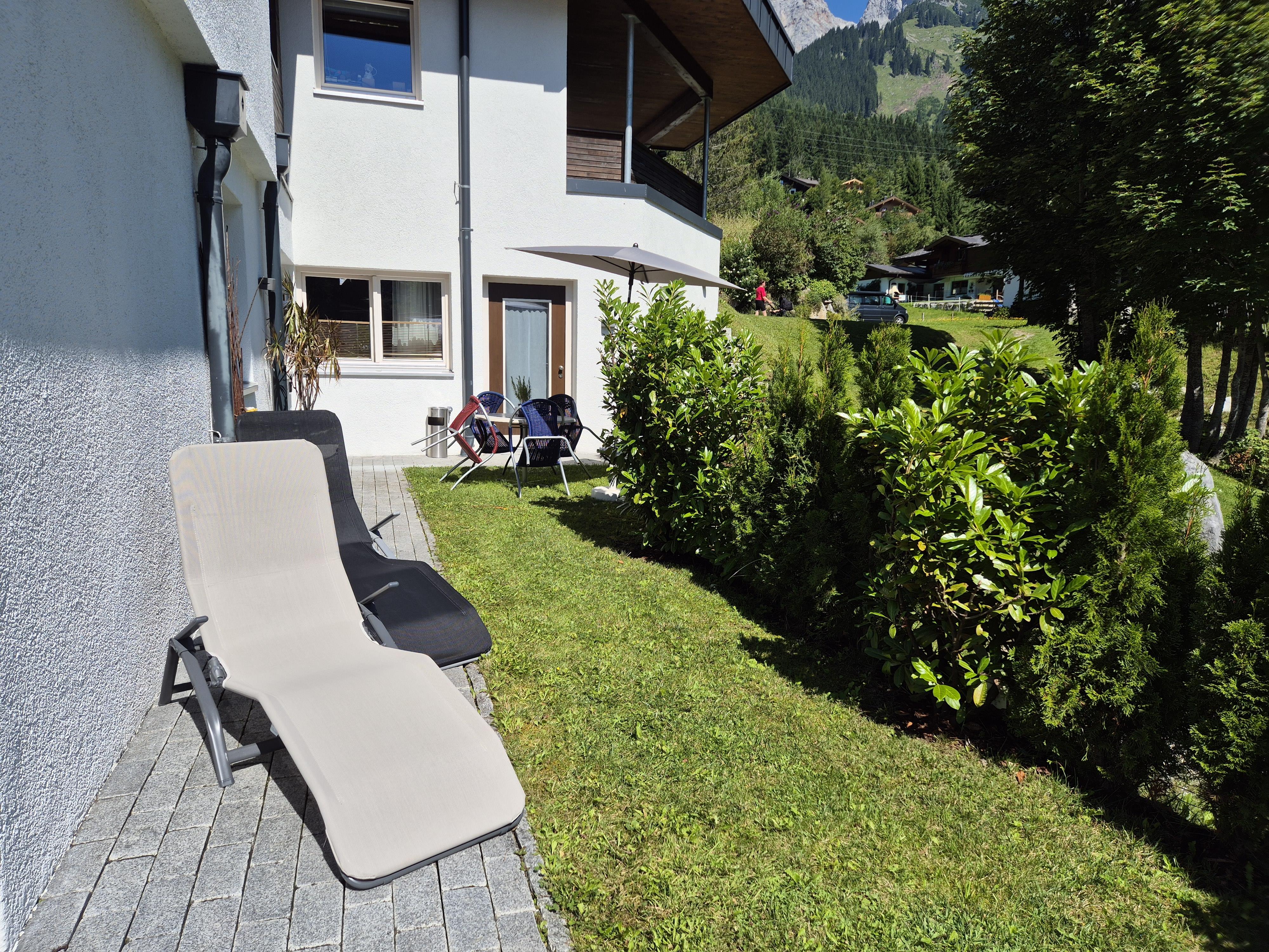 A sunny garden view with loungers and a well-maintained green space. In the background, the mountains and a quiet residential house can be seen.