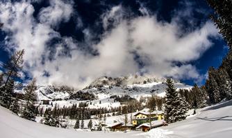 A winter landscape with snow-covered mountains and a clear, blue sky. In the foreground, there are some trees and a cozy building.