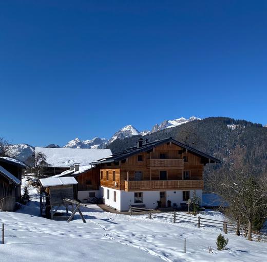 A cozy wooden house in the snow, surrounded by mountains. The clear blue sky creates an idyllic winter landscape.