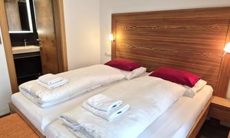A cozy bedroom with a double bed and red pillows. In the background, there is a modern bathroom.